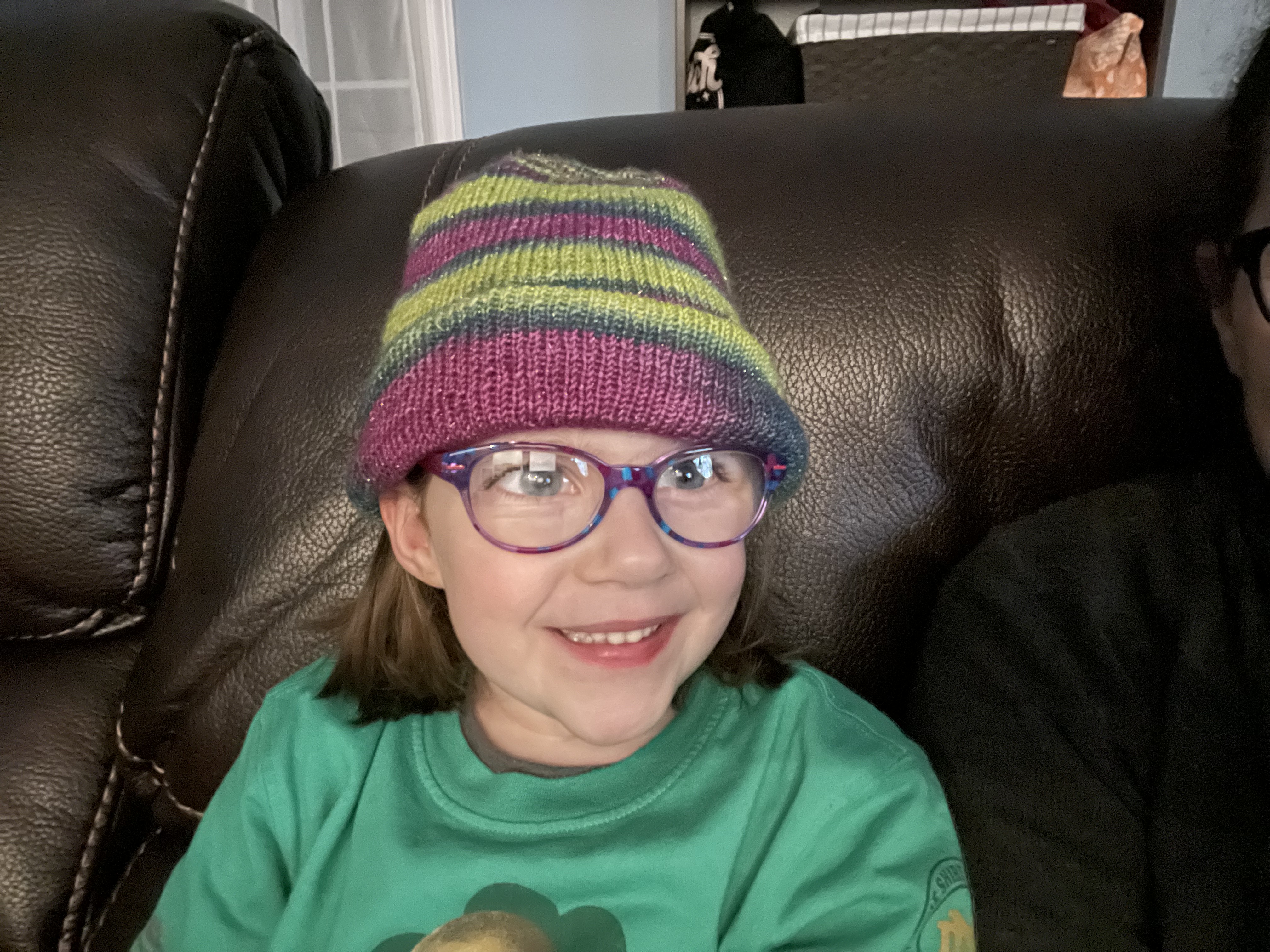A bespectacled child smiles for the camera modeling a green and purple handknitted hat